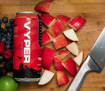 Black and red vyper energy can on cutting board next to knife with blueberries, raspberries, blackberries, an orange, a lime, and chopped apple pieces