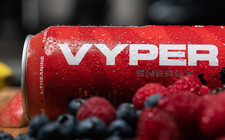 Red and black vyper energy can on a cutting board behind strawberries, rasberries, and blueberries with a banana in the background