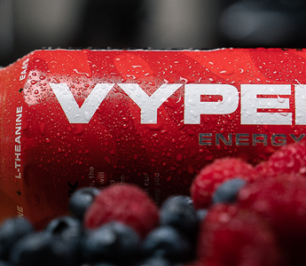 Red and black vyper energy can on a cutting board behind strawberries, rasberries, and blueberries with a banana in the background