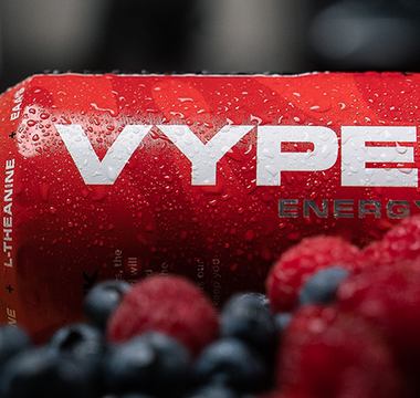 Red and black vyper energy can on a cutting board behind strawberries, rasberries, and blueberries with a banana in the background