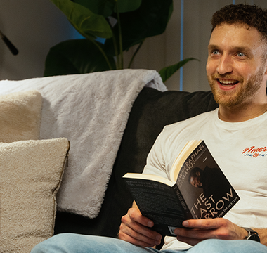 Dayne smiling in his living room sitting on a couch while reading "The Last Arrow" with a Vyper Energy sitting on the table in front of him. 